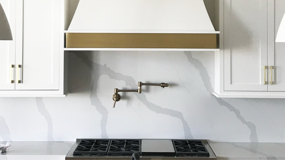 A white rangehood with a golden collar is hung in a white kitchen with a marble backsplash and marble countertops